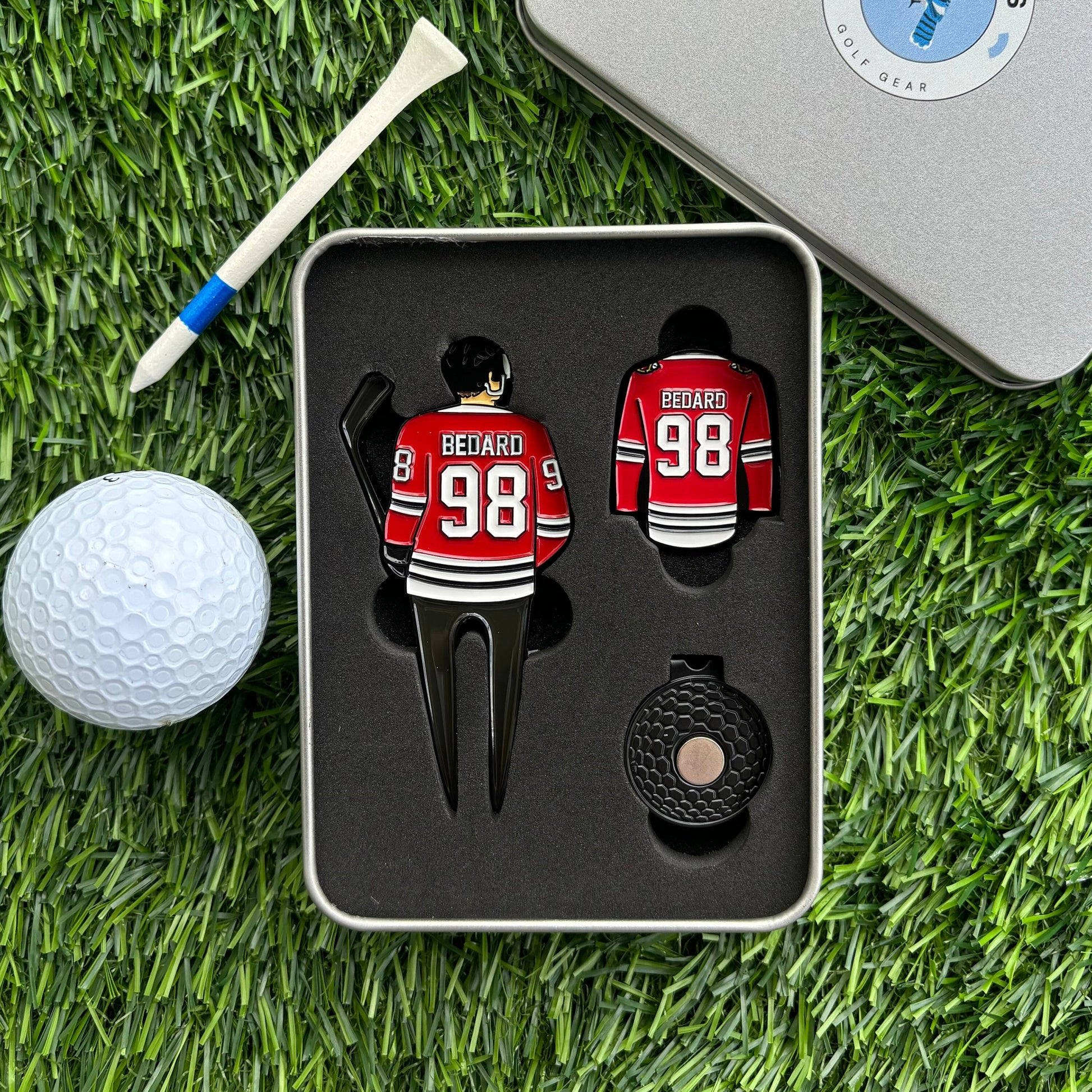 Bedard golf divot tool package with divot tool and golf ball marker in a tin box on grass with tee and golf ball