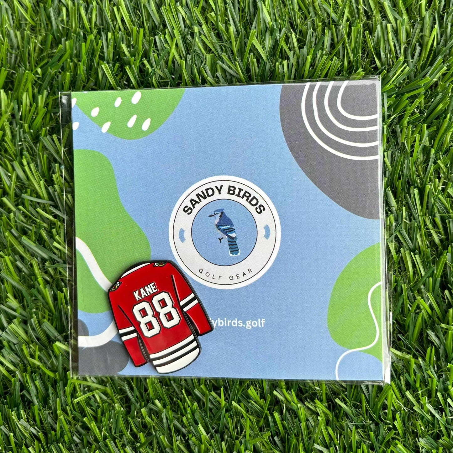 Patrick Kane custom golf ball marker on grass next to Sandy Birds Golf Gear packaging.