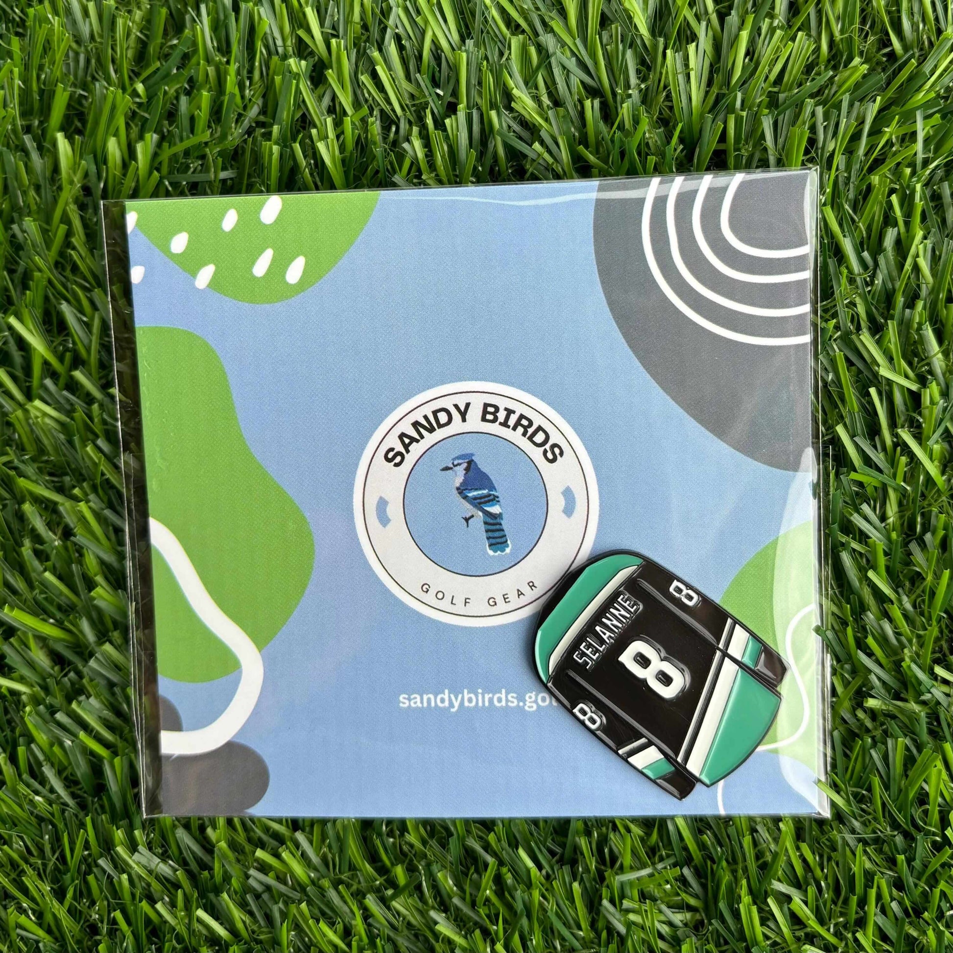 Teemu Selanne golf ball marker on Sandy Birds packaging, placed on green grass.